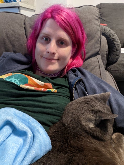 a woman in pink hair smiling at the camera with a small grey cat on her lap